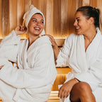 Two women in white robes sitting in a wooden sauna, smiling and laughing.