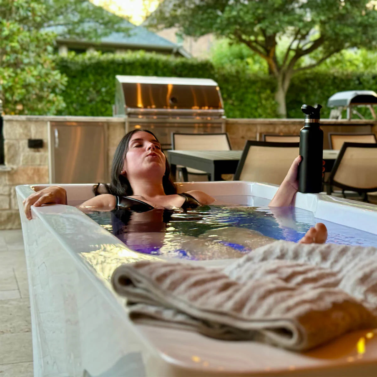 Person relaxing in a hot plunge with a bottle and towel nearby, outdoor setting.