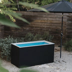 Black rectangular hot tub with blue interior in a garden setting with a wooden fence and umbrella.