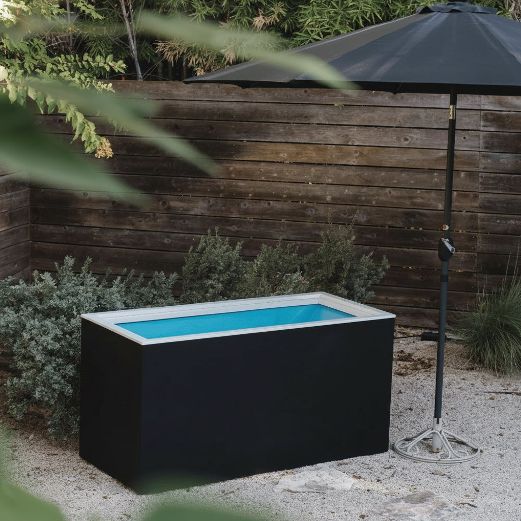 Black rectangular hot tub with blue interior in a garden setting with a wooden fence and umbrella.