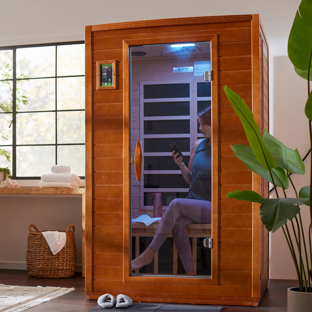 Person using an infrared sauna in a home setting with a plant and window in the background.
