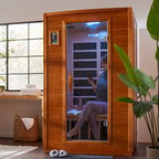 Person using an infrared sauna in a home setting with a plant and window in the background.