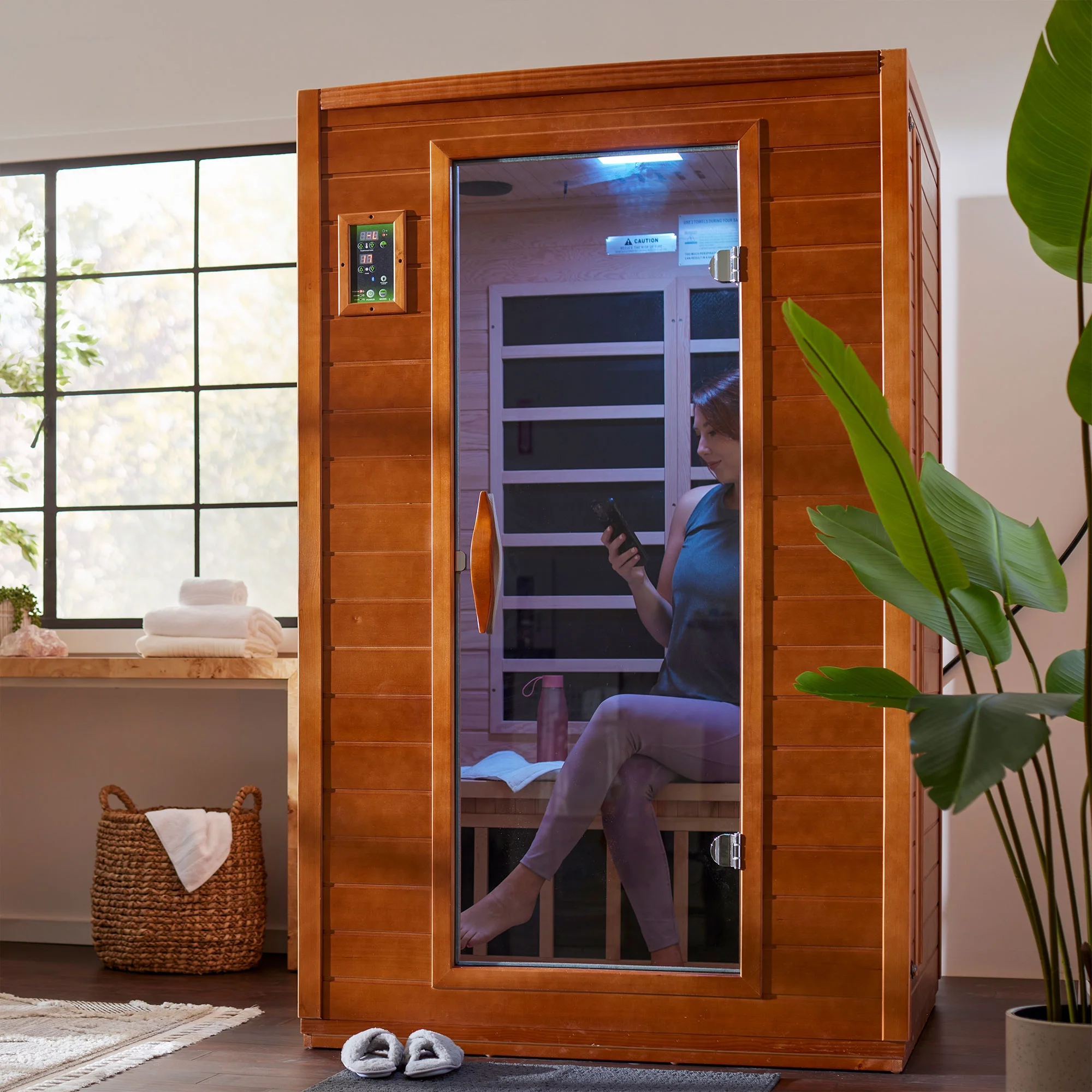 Person using an infrared sauna in a home setting with a plant and window in the background.